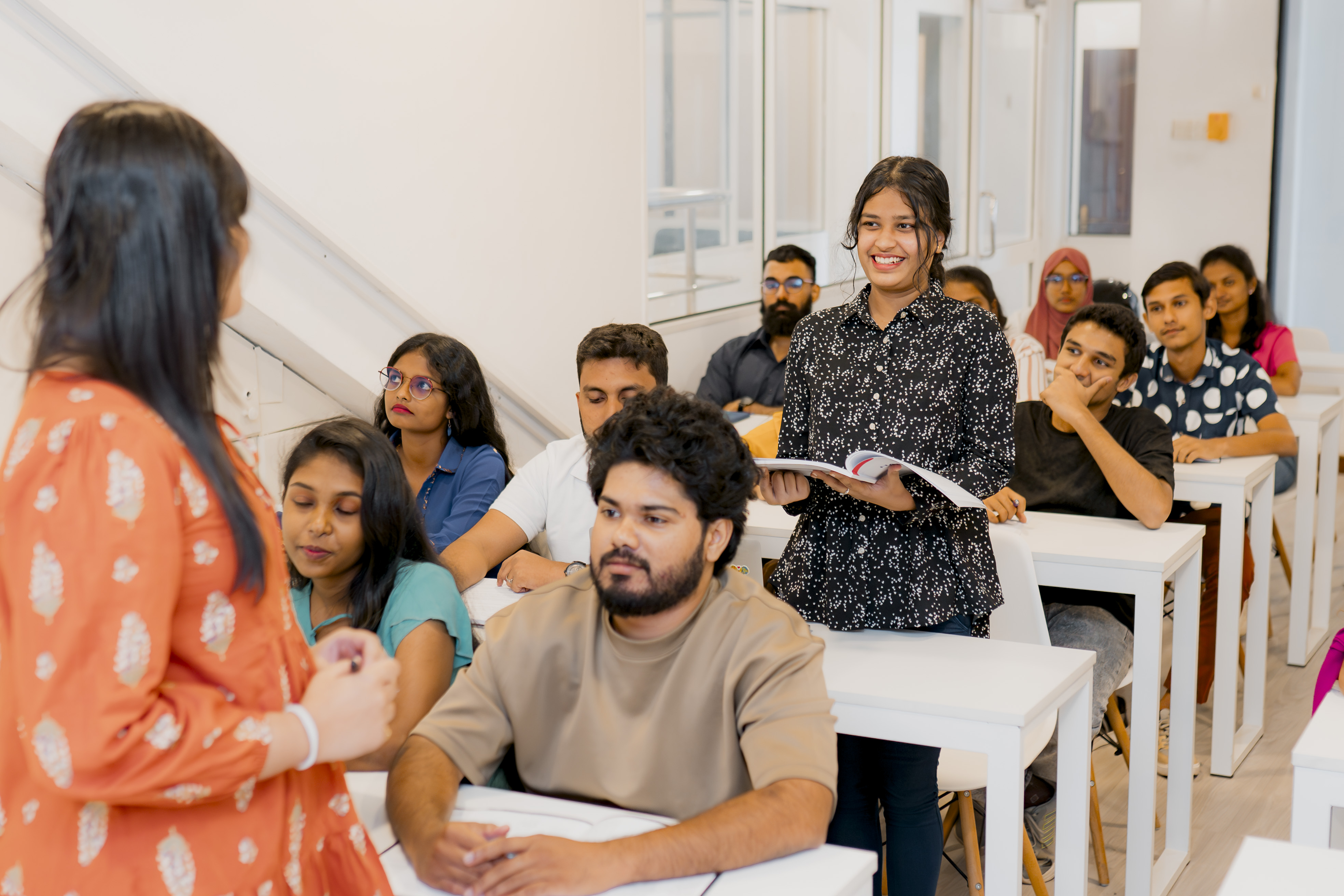 A classroom setting with students attentively listening to a female instructor. One student stands up, smiling and holding a book, while the other students, seated at white desks, exhibit various expressions of interest.